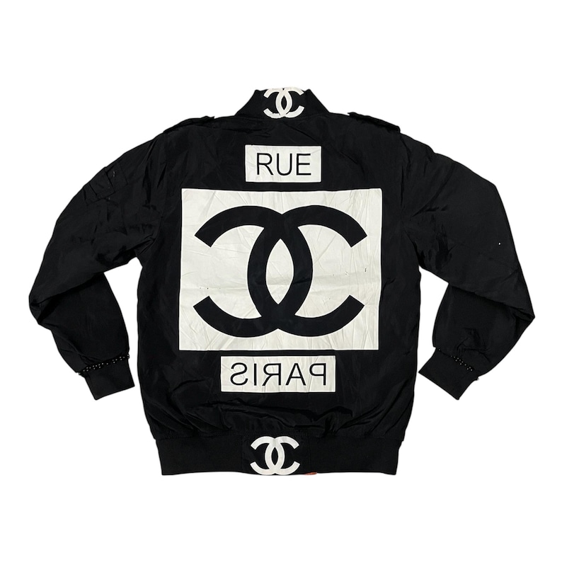 May include: Black bomber jacket with white logo and text. The jacket features a large white square with a double-C logo and the word "RUE" above it. The word "PARIS" is below the logo. The jacket has a stand-up collar.