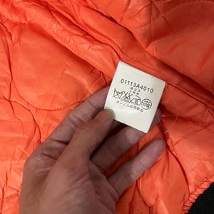 May include: Close-up of a quilted orange jacket interior. A white tag with black text and symbols is visible. The tag includes the numbers "01113A4010", "B23", and "168". The jacket's interior has a diamond pattern.