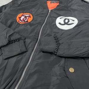 May include: Black bomber jacket with a silver zipper and black ribbed cuffs adorned with small black beads. Features two patches: one orange and black, the other white and black. The jacket has an orange lining.