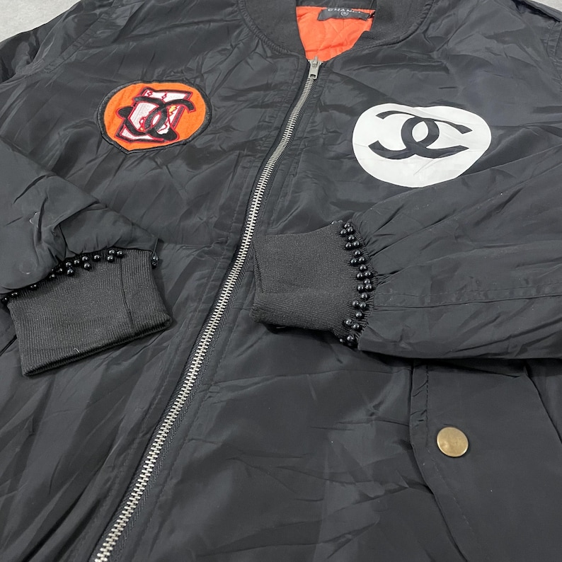 May include: Black bomber jacket with a silver zipper and black ribbed cuffs adorned with small black beads. Features two patches: one orange and black, the other white and black. The jacket has an orange lining.