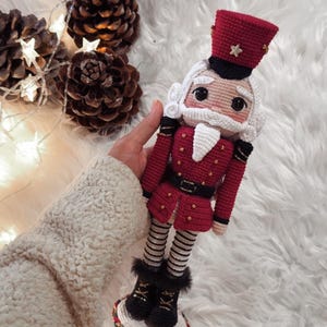 Könnte beinhalten: Handgefertigte gehäkelte Nussknacker-Puppe in roter Uniform mit goldenen Knöpfen, schwarzem Gürtel und hohem roten Hut. Die Puppe hat einen weißen Bart und Haare und steht auf einer weiß-roten Basis. Im Hintergrund sind Tannenzapfen und Lichterketten.