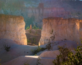 May include: A panoramic view of a canyon with towering sandstone cliffs and a distant mountain range. The canyon floor is covered in sand and sparse vegetation. The sky is a soft blue with a hint of pink.