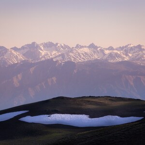 May include: A panoramic view of a mountain range with snow-capped peaks, seen from a grassy hill with patches of snow. The sky is a soft pink and purple.