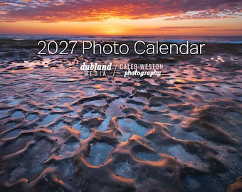 May include: A 2027 photo calendar featuring a landscape photograph of a rocky shoreline at sunset. The image displays tide pools reflecting orange and purple sky colours, with the text 2027 Photo Calendar, dubland MEDIA, and CALEB WESTON photography.