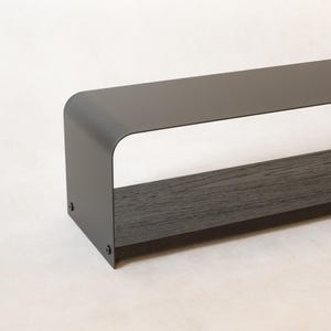Könnte beinhalten: Ein modernes, dunkelgraues Metallregal mit gewölbter Oberseite und dunkler Holzbasis. Das Regal ist rechteckig mit einem hohlen Mittelteil und dient zur Präsentation von Gegenständen. Das minimalistische Design eignet sich für verschiedene Einrichtungsstile.