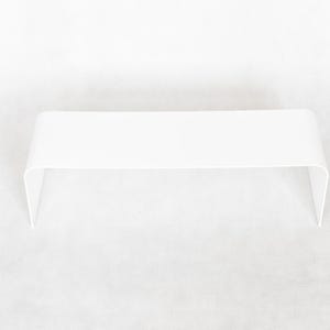 May include: A minimalist, white, modern coffee table with a curved design. The table has a rectangular top and two curved legs. The table is approximately 48 inches long, 16 inches wide, and 14 inches high.