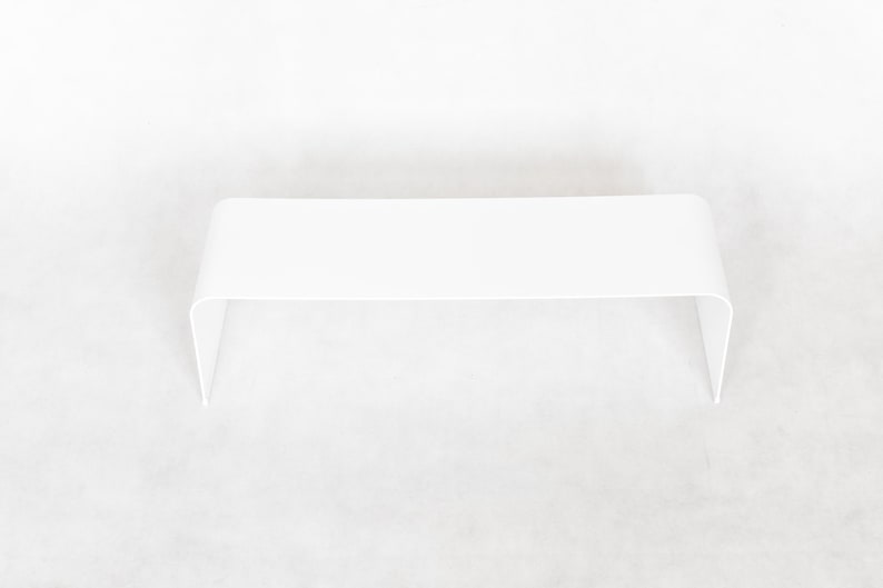 May include: A minimalist, white, modern coffee table with a curved design. The table has a rectangular top and two curved legs. The table is approximately 48 inches long, 16 inches wide, and 14 inches high.