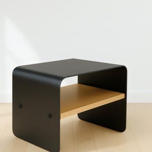 A black metal nightstand with a light wood shelf, shown at an angle on a light wood floor against a white wall.