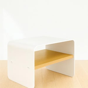 A white metal nightstand with a light wood shelf, shown at an angle on a light wood floor against a white wall.