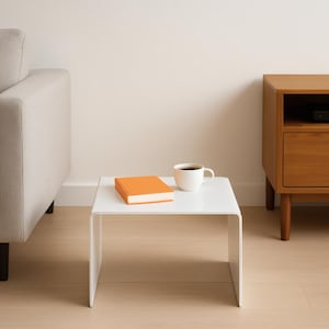 A white minimalist coffee table with an orange book and a white mug placed on it, positioned next to a gray sofa.