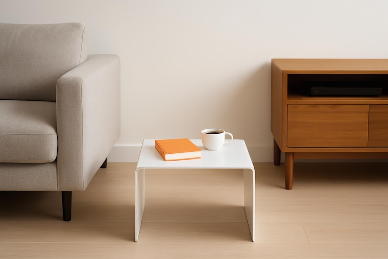 A white minimalist coffee table with an orange book and a white mug placed on it, positioned next to a gray sofa.