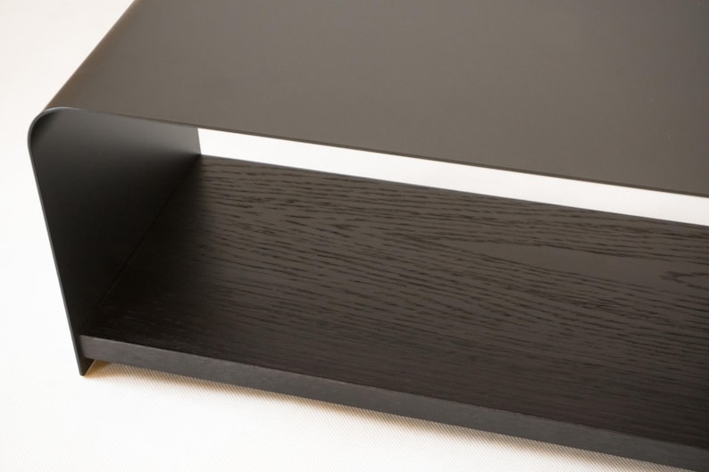 May include: A contemporary coffee table with a dark, matte finish. The table has a rectangular shape with a solid, dark wood grain shelf and a sleek, curved metal top. The design is minimalist and modern, suitable for a living room.