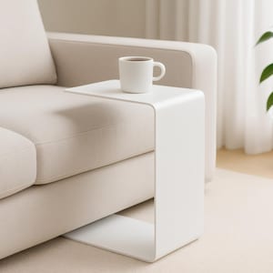 A white C-shaped metal side table is positioned next to a light beige sofa, holding a white mug with a beverage.