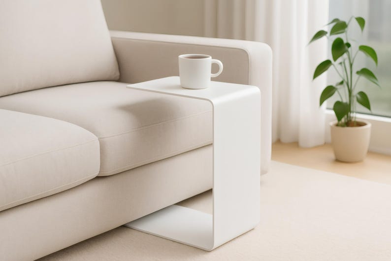 A white C-shaped metal side table is positioned next to a light beige sofa, holding a white mug with a beverage.