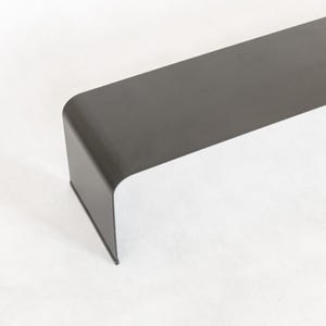 May include: A long, dark gray metal bench with a curved edge. The bench is made of a single piece of metal and has a modern, minimalist design.