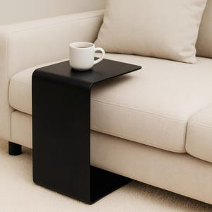 A black C-shaped metal side table is positioned next to a light beige sofa, holding a white mug with a beverage.