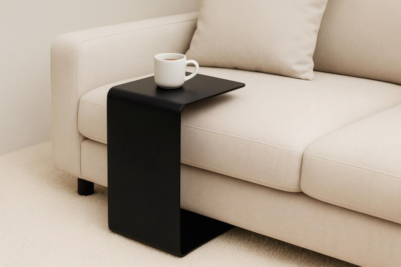 A black C-shaped metal side table is positioned next to a light beige sofa, holding a white mug with a beverage.