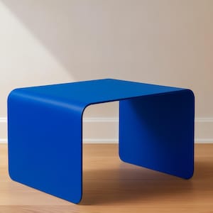 An ultramarine blue curved metal side table sitting on a light wood floor next to a white wall.