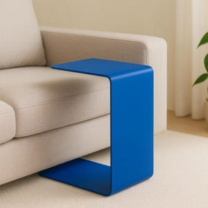 A blue C-shaped metal side table is placed with its base under a light beige sofa, extending over the seat.