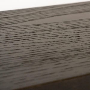 May include: Close-up of a section of dark brown wood with a visible grain pattern. The wood appears to be part of a larger structure, possibly a table or a piece of furniture. The surface is smooth and has a slight sheen.