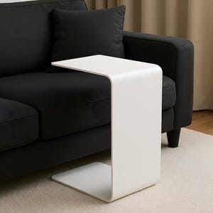 A white C-shaped metal side table is placed with its base under a black sofa, extending over the seat.