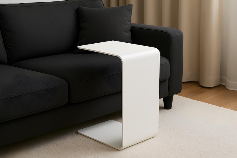 A white C-shaped metal side table is placed with its base under a black sofa, extending over the seat.