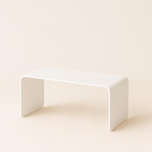 A white minimalist metal coffee table with rounded edges on a pale surface against a neutral background.