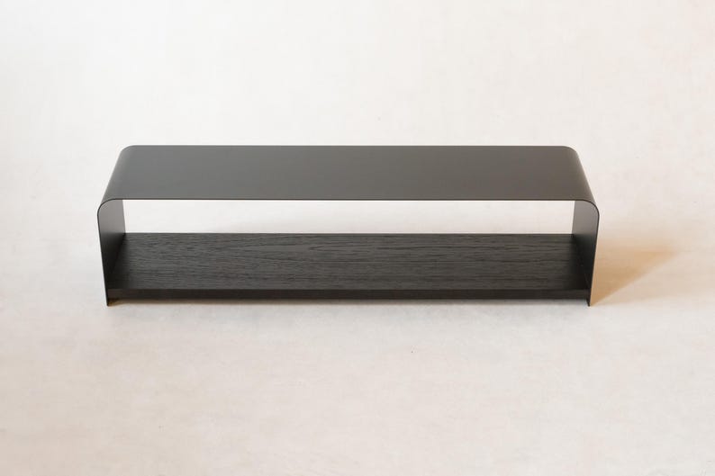 May include: A contemporary black shelf with a curved top and a wooden base. The shelf is made of metal and wood. It is designed to be mounted on a wall or placed on a surface. The shelf is approximately 60 cm long.