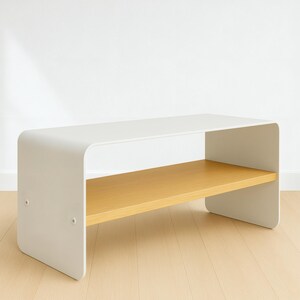 A white metal bench with rounded corners and a light wood shelf, shown at an angle on a light wood floor against a white wall.