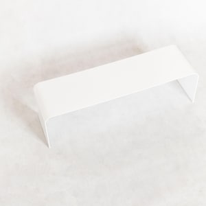 May include: A minimalist, white, rectangular shelf with rounded corners and a curved, open design. The shelf is made of a solid, opaque material and is elevated, creating a modern aesthetic.