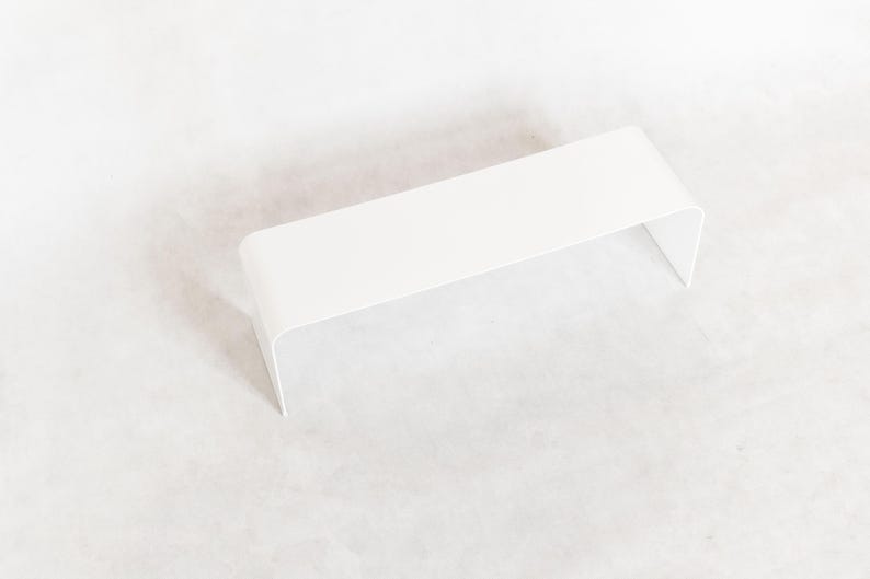 May include: A minimalist, white, rectangular shelf with rounded corners and a curved, open design. The shelf is made of a solid, opaque material and is elevated, creating a modern aesthetic.