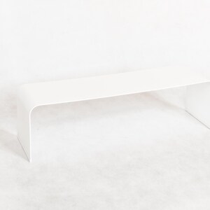 May include: A modern, minimalist white bench with a curved design. The bench is approximately 36 inches long and 12 inches wide, with a seamless, continuous surface. The legs are integrated into the design, creating a clean, contemporary aesthetic.