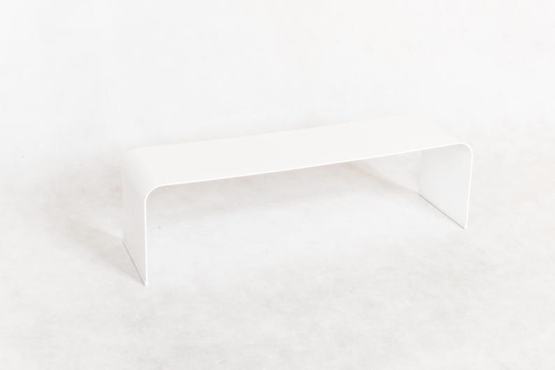 May include: A modern, minimalist white bench with a curved design. The bench is approximately 36 inches long and 12 inches wide, with a seamless, continuous surface. The legs are integrated into the design, creating a clean, contemporary aesthetic.