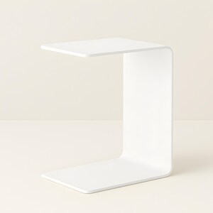 A white C-shaped metal side table is shown from a slight angle against a light beige background.