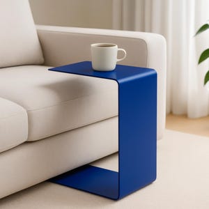 A blue C-shaped metal side table is positioned next to a light beige sofa, holding a white mug with a beverage.
