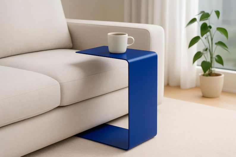 A blue C-shaped metal side table is positioned next to a light beige sofa, holding a white mug with a beverage.