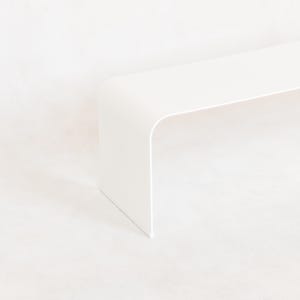 May include: A minimalist, white, modern bench with a curved design. The bench has a rectangular top and a single, curved leg. The bench is made of a solid, opaque material and is set against a white background.