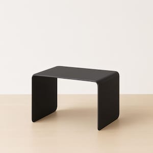 A black minimalist bent metal side table on a light wood floor against a white wall.