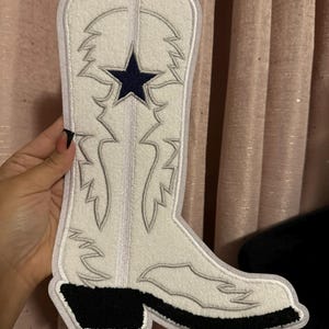 May include: A white cowboy boot-shaped patch with a navy blue star and gray stitching. The boot has a black heel and sole. The patch is made of a soft, felt-like material.
