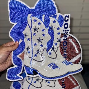 May include: A sequined decoration in the shape of a cowboy boot, bow, and football. The boot is white with blue accents and star details. The word "COWBOYS" is written vertically. The football is brown and red.