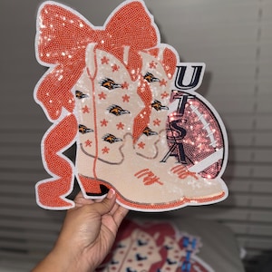 May include: A decorative cutout featuring a pair of sequined cowboy boots, a football, and a bow. The boots are cream-colored with orange accents and roadrunner bird designs. The football is pink and the bow is orange.