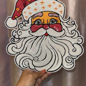 May include: A large, white, sparkly, sequined Santa Claus face patch with a red hat and a white beard. The patch is perfect for adding a festive touch to any project.