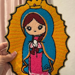 May include: A colorful embroidered patch featuring a cartoon depiction of the Virgin Mary. The patch is oval-shaped and has a gold border. The Virgin Mary is wearing a blue robe and a pink dress. She has a crown on her head and is holding a white flower.