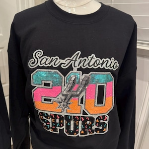 May include: Black sweatshirt featuring the text "San Antonio" in silver script above a sequined "210" and "SPURS" logo. The "210" is in teal, pink, and orange. The Spurs logo is in silver.