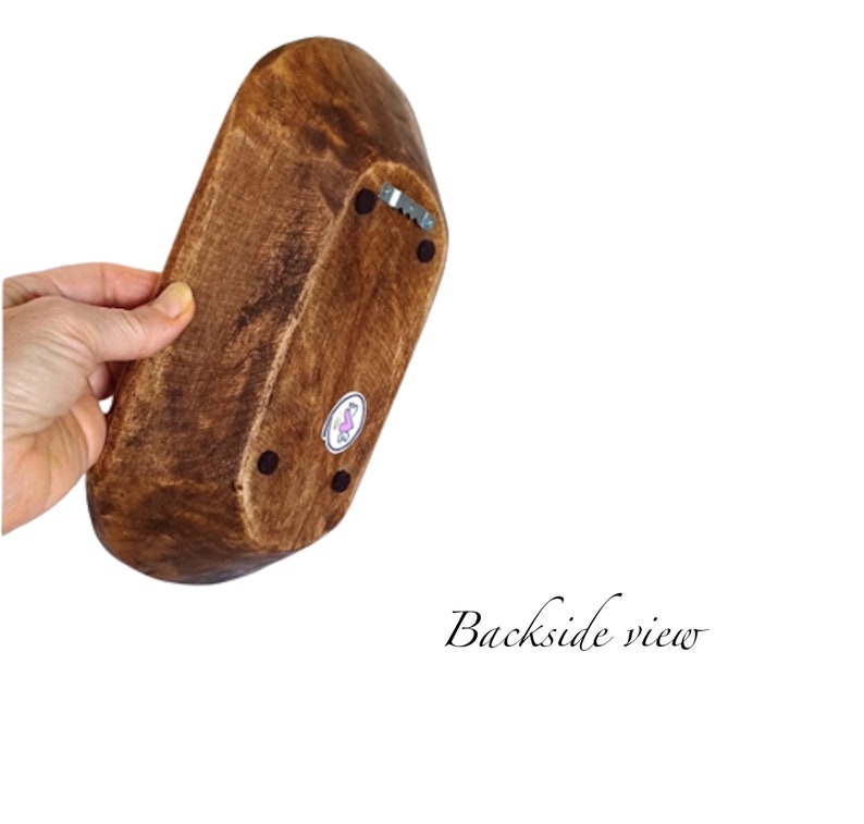 May include: A wooden bowl with a dark brown stain. The bowl has a rounded oval shape and is shown from the bottom. The bowl has three small black dots on the bottom and a small silver metal hook.