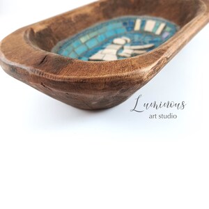 May include: A wooden dough bowl with a blue and white mosaic design featuring a stylised angel figure. The bowl is made of natural wood and has a rustic, handcrafted look.