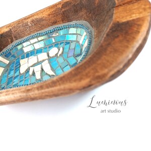 May include: A wooden bowl with a blue mosaic design inside. The bowl is oval shaped and has a dark brown wood finish. The mosaic design features a variety of blue and white tiles. The text "Luminous art studio" is written in cursive on the bottom right corner of the bowl.