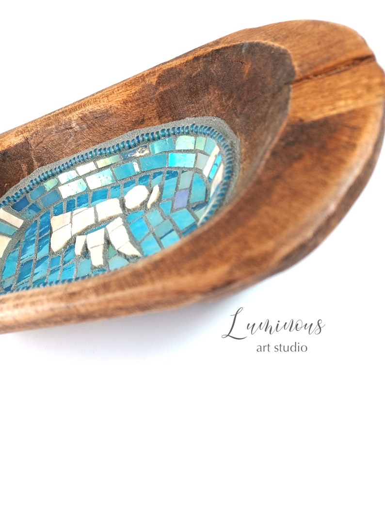 May include: A wooden bowl with a blue mosaic design inside. The bowl is oval shaped and has a dark brown wood finish. The mosaic design features a variety of blue and white tiles. The text "Luminous art studio" is written in cursive on the bottom right corner of the bowl.