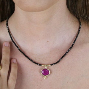 May include: A black beaded necklace with a gold pendant featuring a large, faceted, pink gemstone. The pendant is shaped like a pomegranate.