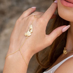 May include: A woman wearing a white dress with gold jewelry. She has a gold chain bracelet on her left wrist with a floral design, a gold necklace with a floral design, and a gold ear cuff on her right ear.